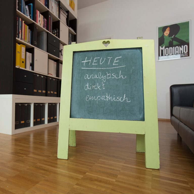Rustikaler Aufsteller aus Holz mit Kreidefläche, dieser steht auf Parkett in einem wertigen Büro mit seitlicher Regalwand.