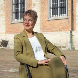 Sandra Kroning auf Stuhl sitzend, Kopfsteinpflaster und Gebäude im Hintergrund mit Backsteinziegeln